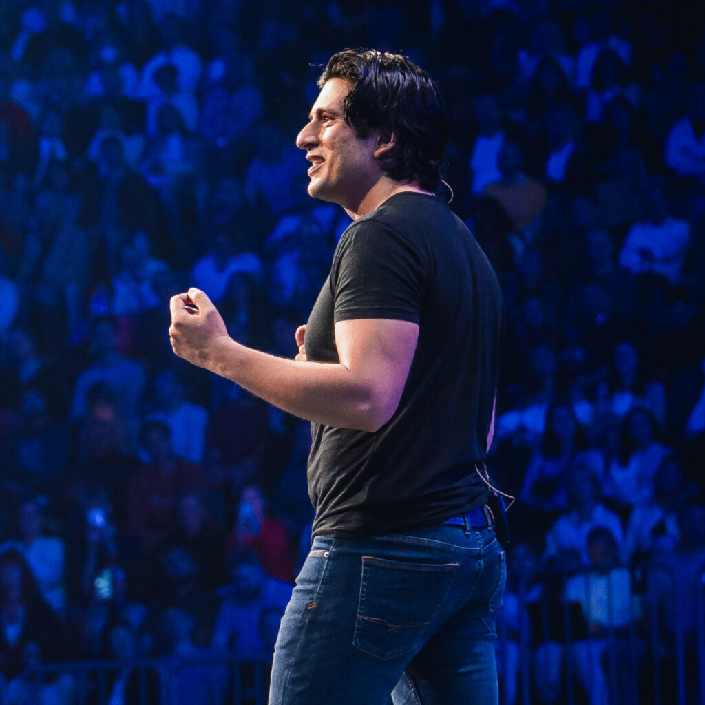 ⁠⁠Francisco Medina in schwarzem T-Shirt und Jeans steht im Profil auf einer Bühne. Er gestikuliert mit geballter Faust und lächelt. Im Hintergrund ist ein großes Publikum unter blauer Beleuchtung zu sehen.
