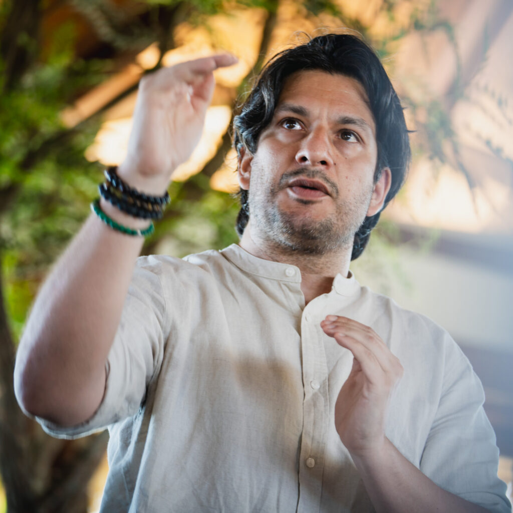 ⁠⁠⁠⁠⁠⁠Francisco Medina in weißem Hemd mit Stehkragen gestikuliert während eines Vortrags. Im Hintergrund sind unscharfe grüne Pflanzen und helles Licht zu erkennen.