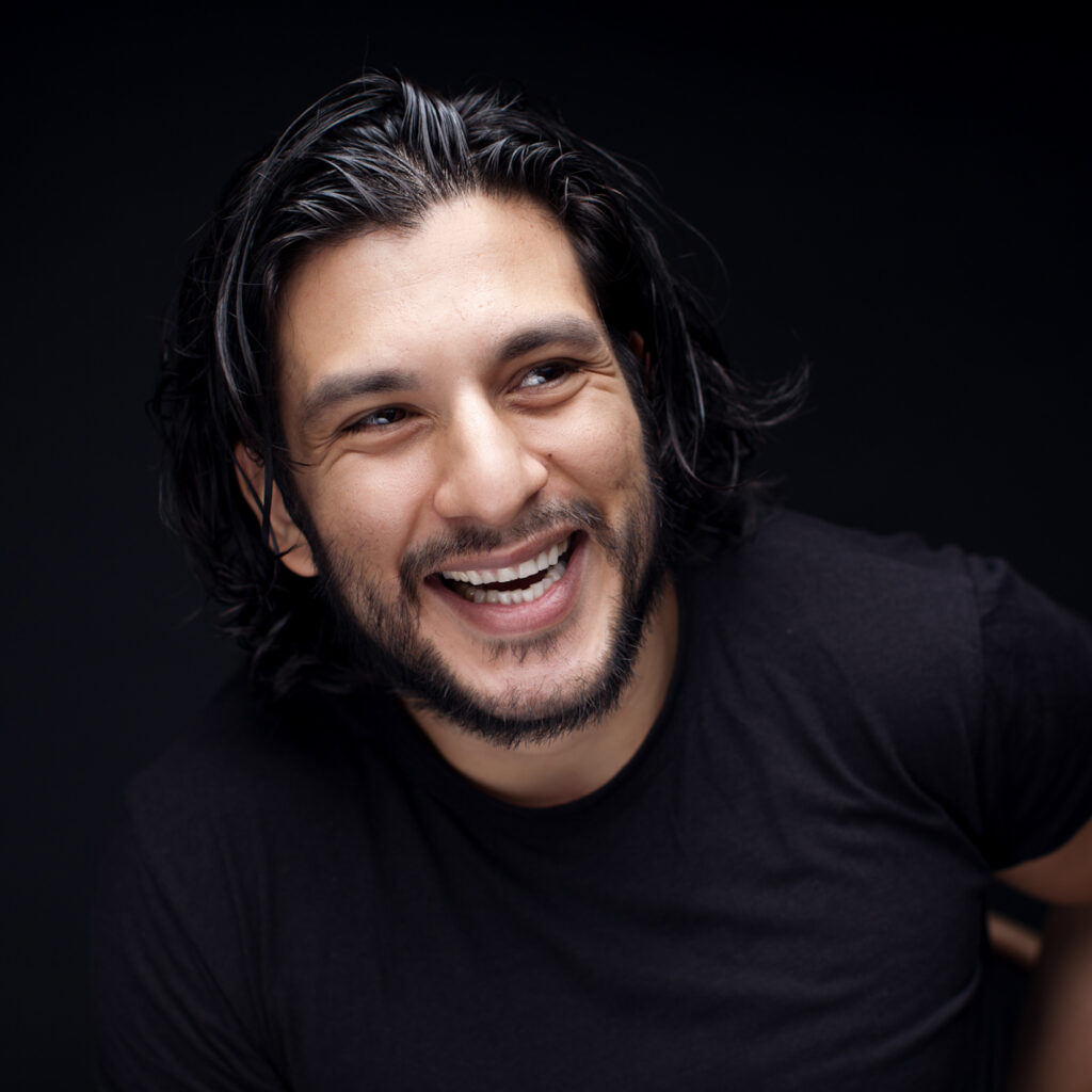 Portrait von ⁠⁠⁠⁠Francisco Medina, der nach vorne gelehnt und oben blickend herzlich lacht, in schwarzem T-Shirt vor schwarzem Hintergrund. Es ist ein Foto-Shooting.
